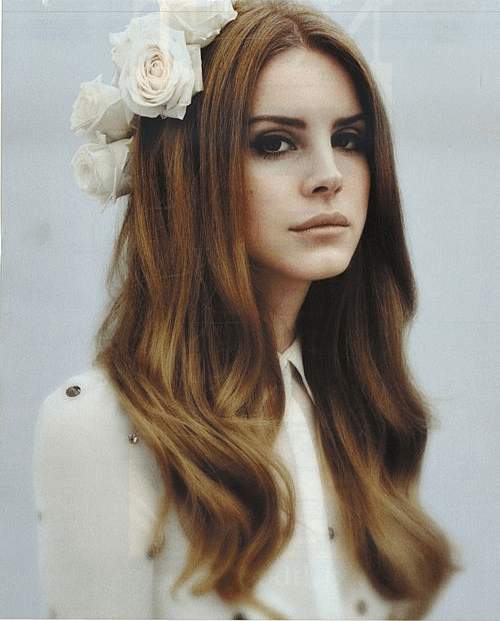 Lana Del Rey stares blankly at a spot behind the camera/ She's wearing white, with a white flower in her hair. He hair is brown and curled and she is sat against an off-white background.
