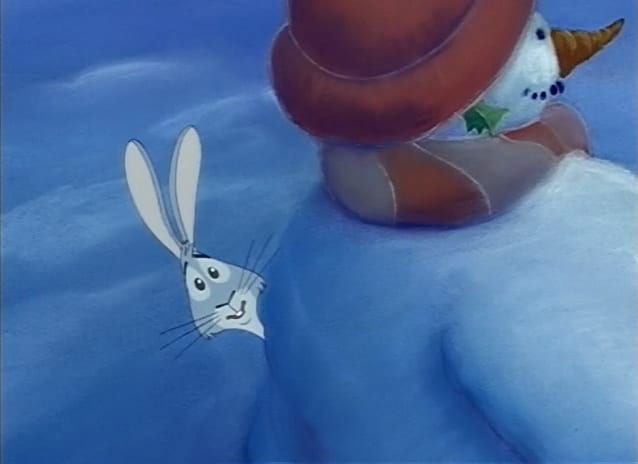 An animated grey rabbit peeking out from behind a snowman stood sideways with a reddy orange hat and scarf.