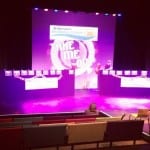 A stage with two stands with women's names on which is empty. A massive sign is on a purple and pink screen saying "St Barnabas Lincolnshire Hospice" and "Take Me Out"